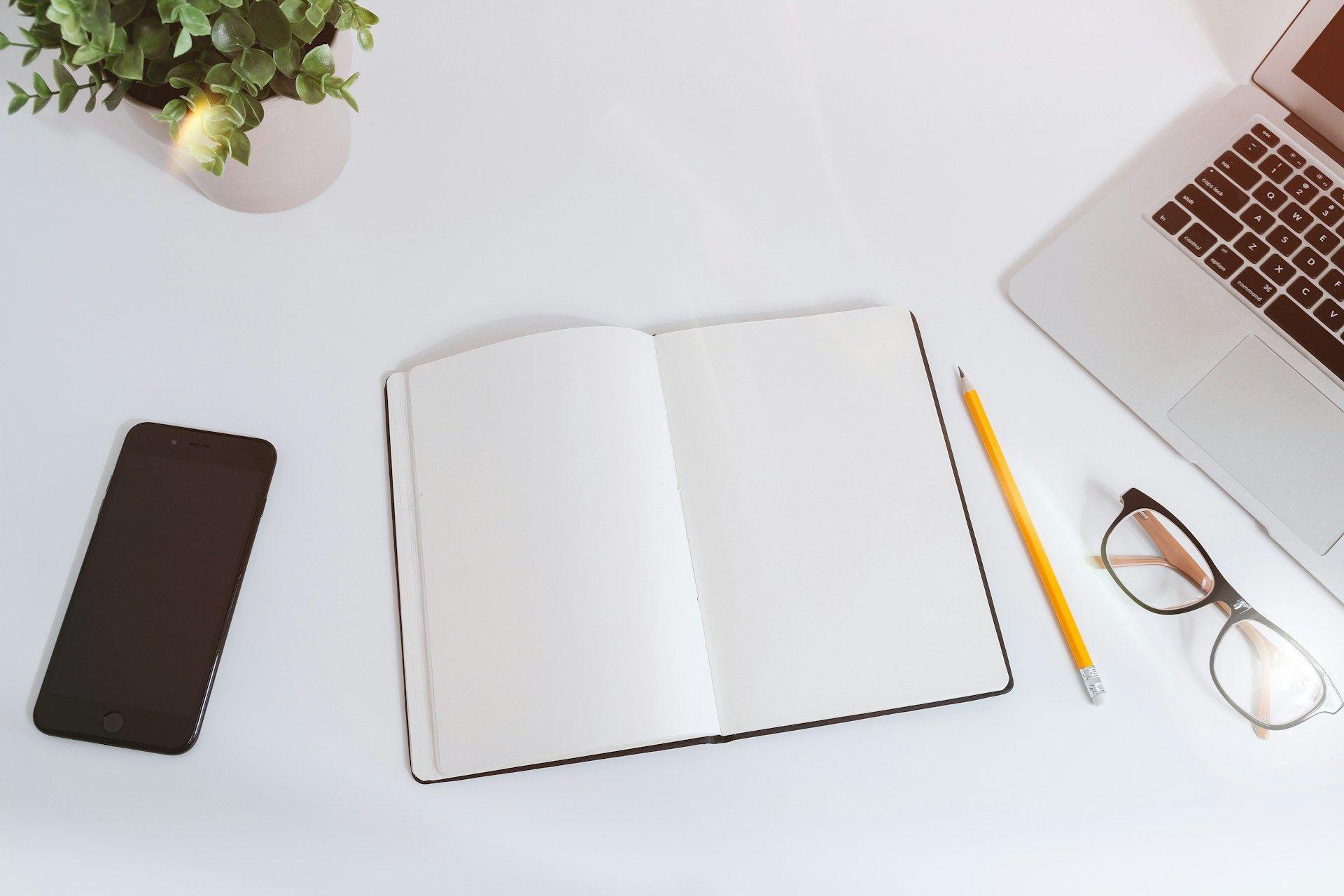 um caderno, telefone, óculos e uma planta em uma mesa branca