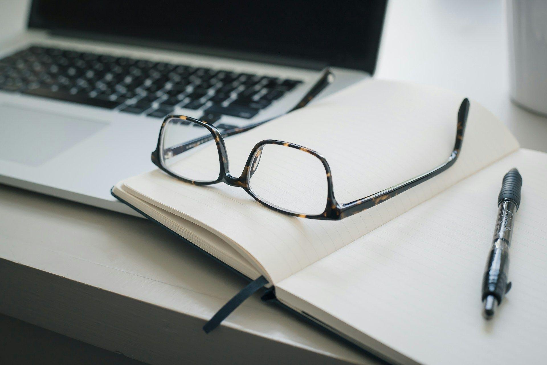 a notebook, glasses and a laptop on a desk