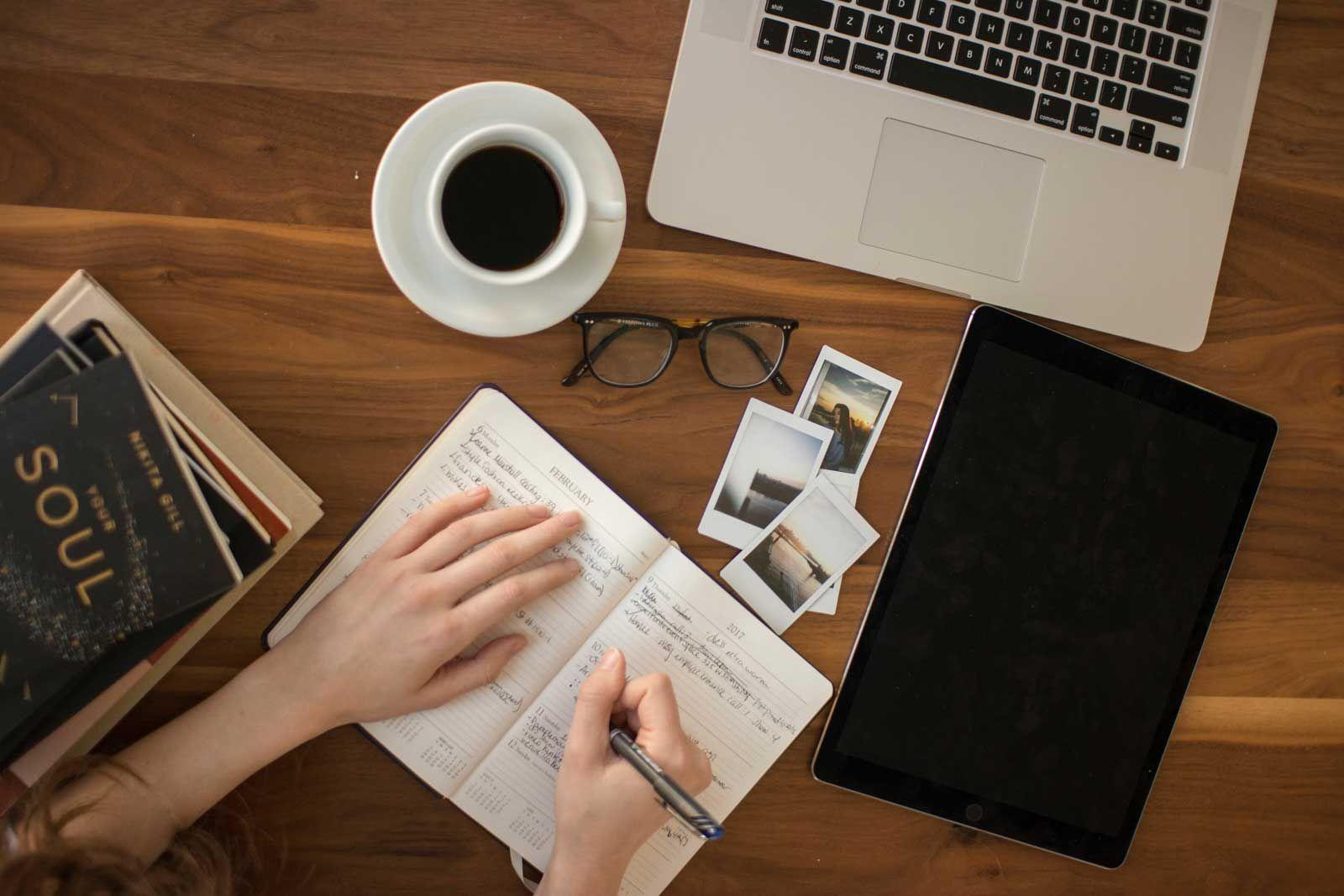 een vrouw die in een notitieboekje schrijft aan een houten tafel met een kop koffie