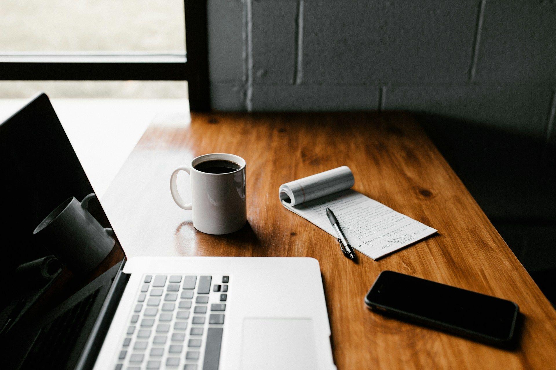 um laptop em uma mesa de madeira ao lado de um caderno e uma xícara de café