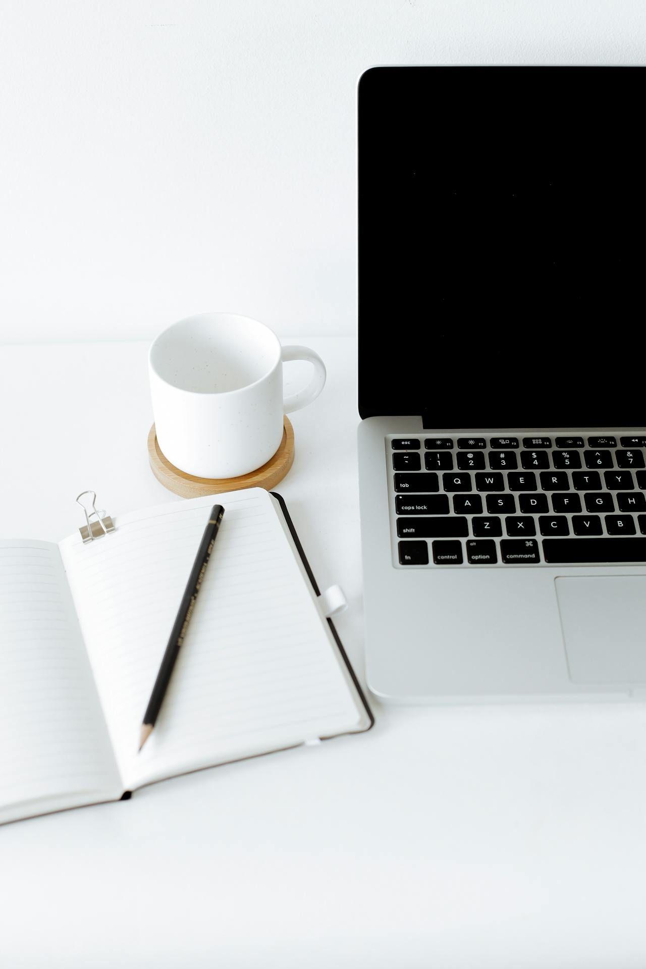 a laptop, a notebook, and a cup of coffee on a white table