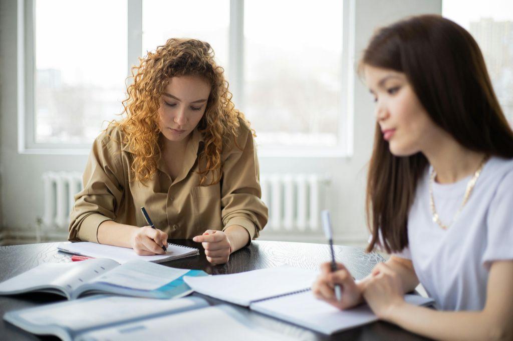 dos mujeres sentadas en una mesa y escribiendo en cuadernos